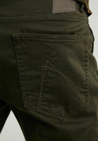 Olivgröna byxor med en distinkt vinklad bakfickedesign, med kontrasterande sömmar och en läderlogotyp på midjebandet.