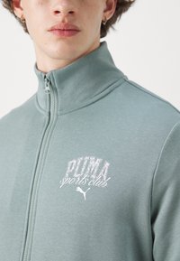 Jasnozielona kurtka zapinana na zamek, wykonana z miękkiego materiału, z wysokim kołnierzem i białym nadrukowanym logo z przodu.