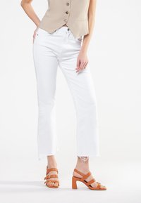 Hvide jeans med frynset kant parret med en beige cropped vest. Brune læderhæle sandaler med flere stropper. Rent, minimalistisk design.