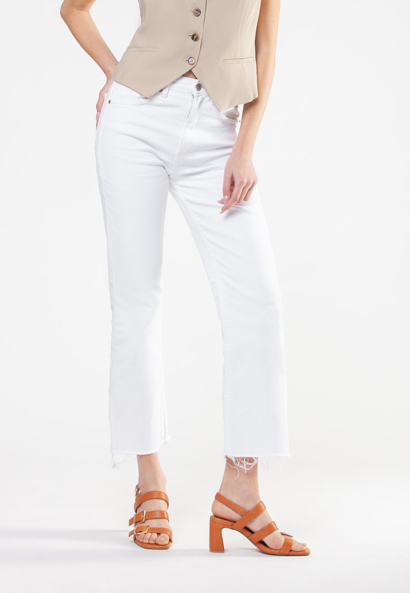 Hvide jeans med frynset kant parret med en beige cropped vest. Brune læderhæle sandaler med flere stropper. Rent, minimalistisk design.
