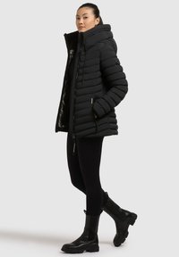 Frau steht da und trägt eine schwarze gefütterte Jacke mit Kapuze, schwarze Leggings und schwarze klobige Stiefel vor einem schlichten hellen Hintergrund.