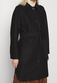 Cappotto di lana nera con colletto, chiusura a bottoni, due tasche e cintura in tessuto. Il design presenta una silhouette dritta e spacchi laterali.