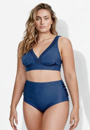 Donna con capelli lunghi e mossi che indossa un costume da bagno due pezzi blu con bordo arricciato sul top e slip a vita alta su sfondo bianco.