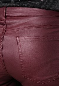 Burgundské přiléhavé kalhoty s hladkou texturou, které mají zadní kapsy a jemné švy. Materiál vypadá jako pružný.