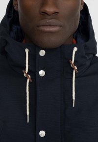 Navy blue jacket with a hood, featuring cream drawstrings, silver snaps, and a textured fabric. Includes a visible inner lining.
