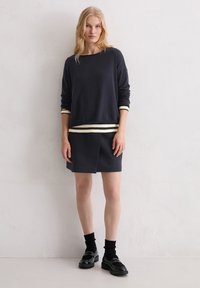 Donkerblauwe trui met gestreepte witte en crèmekleurige boorden, gecombineerd met een bijpassende donkerblauwe rok met een plooi aan de voorkant. Zwarte schoenen en sokken.