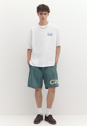 Jeune homme portant un t-shirt blanc oversize, un short vert à carreaux avec le texte "City", des chaussettes blanches et des chaussures marron foncé, debout les mains dans les poches.