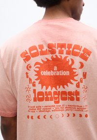 Άτομο που φοράει ροζ μπλουζάκι με πορτοκαλί γραφικά ήλιου και φεγγαριού και κείμενο σχετικά με εορτασμό ηλιοστασίου στην πλάτη.