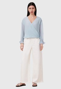 Hellblaues crinkle Wrap-Oberteil mit langen Ärmeln, kombiniert mit lockeren weißen Leinenhosen und flachen silbernen Sandalen. Minimalistisches Design und Textur.