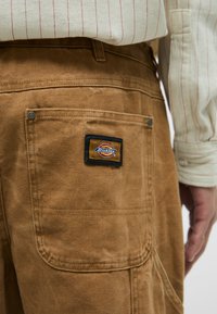 Καφέ παντελόνι από καμβά με μια πίσω τσέπη που έχει μια μαύρη παστούρα με την ετικέτα "Dickies" και μεταλλικό κούμπωμα. Υφασμάτινος ιστό με διακριτική ραφή.