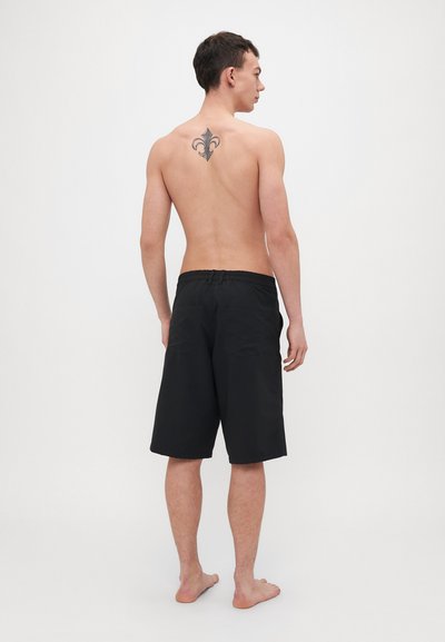 Černé plavky s elastickým páskem, obsahující dvě zadní kapsy a hladkou texturu. Model je zobrazen zezadu.