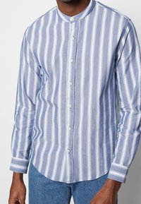 Knappt upp-skjorta med blå och vita vertikala ränder. Tillverkad av lättviktig tyg, med en rundad krage och förlängda manschetter.