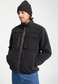 Homme portant une veste en polaire grise foncée avec des poches poitrine zippées et à rabat, un jean bleu et un bonnet en tricot noir, se tenant avec les mains dans les poches.