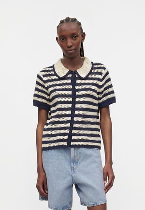 Νεαρή γυναίκα φοράει μία ριγέ πλεκτή ζακέτα σε ναυτικό μπλε και κρέμ χρώμα με γιακά Peter Pan και ανοιχτό μπλε τζιν σορτς.