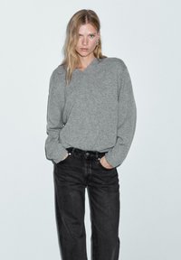 Maglione grigio lavorato a maglia con scollo a V e maniche lunghe, abbinato a jeans neri in denim. Il materiale appare morbido e leggermente strutturato.