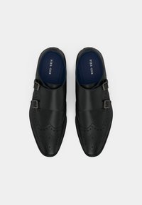 Černé kožené boty s dvojitými přezkami, špičatou špičkou, broguovými detaily a hladkou strukturou. Podšívka s viditelným logem značky.