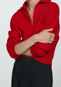 Persona con le braccia incrociate, che indossa un maglione rosso a coste con mezza cerniera e pantaloni scuri a vita alta su uno sfondo neutro.