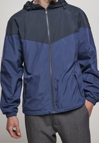 Marinblå och mörkblå huva vindjacka med elastiska ärmslut, framsidan med dragkedjestängning och sidofickor. Slätt, lättviktigt tyg med färgblockering.