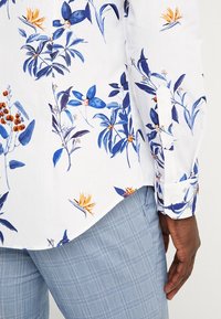 Biała koszula w niebieski kwiatowy wzór, z długimi rękawami i zapinaną mankietą, w zestawie z jasnoniebieskimi spodniami w kratę.