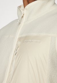 Bela flis in gladek fabric vest z visokim ovratnikom, zadrgo in logotipom "PeakPerformance" na prsih. Teksturiran in plasten dizajn.