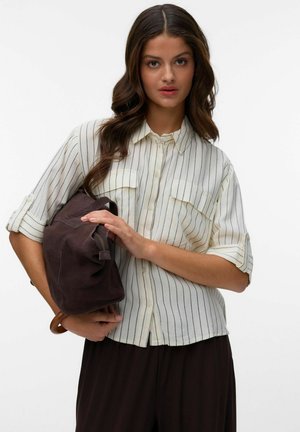 Kobieta o długich, falujących włosach, ubrana w białą koszulę z cienkimi czarnymi paskami, trzymająca ciemnobrązową skórzaną torbę na jednolitym tle.