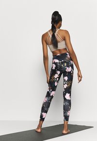 Legginsy w kwiatowy wzór w czerni z różowymi i białymi kwiatami, połączone z beżowym sportowym biustonoszem na ramiączkach. Modelka stoi na macie do jogi.