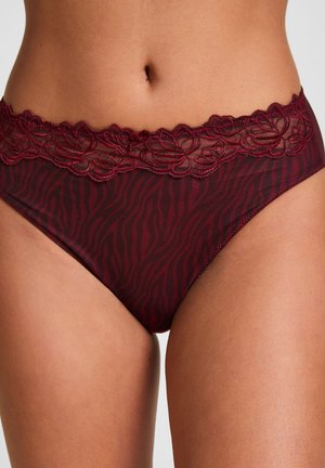 Femme portant un sous-vêtement rouge foncé à motifs avec bordure en dentelle sous le nombril sur un fond uni.