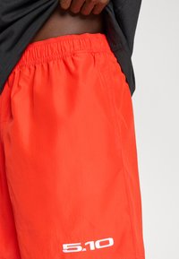 Φωτεινά πορτοκαλί σορτς από ελαφρύ ύφασμα, με ελαστική μέση και λευκό λογότυπο στο αριστερό τελείωμα. Ομαλή υφή.