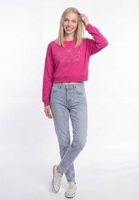 Jonge vrouw die lacht, gekleed in een roze sweatshirt met de tekst "Happy Girls", lichtblauwe spijkerbroek en witte sneakers, staand met één hand op haar hoofd.