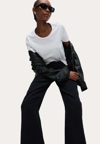 Witte T-shirt, zwarte leren jas met oversized mouwen en zwarte brede broek. Model draagt donkere zonnebril. Effen achtergrond.