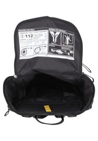 Sac à dos noir avec une ouverture à zip, présentant un intérieur vide et une étiquette d'instruction d'aide d'urgence imprimée à l'arrière.