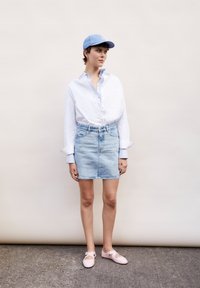 Casquette bleu clair, chemise blanche à boutons, jupe en denim clair et ballerines roses avec des découpes. Fond uni avec un sol texturé.