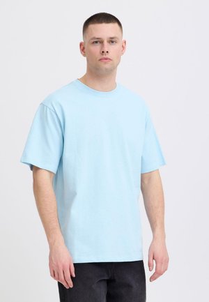 Jeune homme aux cheveux courts portant un t-shirt bleu clair à manches courtes et un pantalon foncé, debout devant un fond blanc uni.