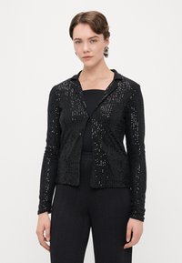 Czarna długa kurtka z długim rękawem, ozdobiona cekinami, z wyciętym kołnierzem i zapięciem z przodu, w zestawieniu z czarnym topem i fakturowanymi czarnymi spodniami.