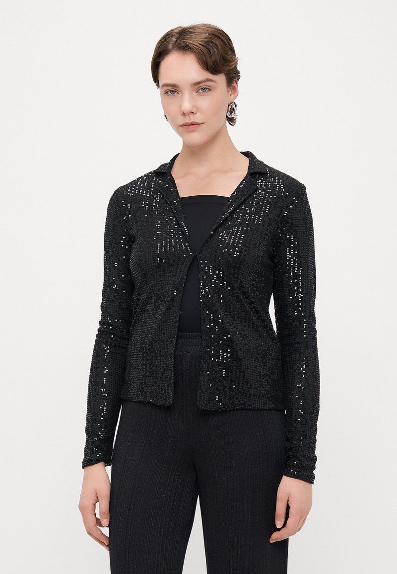 Czarna długa kurtka z długim rękawem, ozdobiona cekinami, z wyciętym kołnierzem i zapięciem z przodu, w zestawieniu z czarnym topem i fakturowanymi czarnymi spodniami.