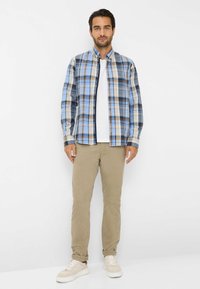 Geruite button-up shirt in blauw, beige en bruin, gedragen over een wit t-shirt, gecombineerd met beige broek en lichte sneakers. Minimalistisch ontwerp.