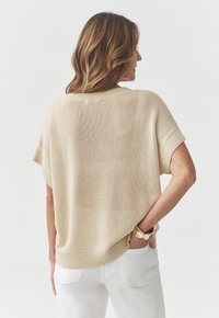 Maglione beige lavorato a maglia con maniche corte e vestibilità ampia. Presenta un colletto ampio e un orlo a coste, abbinato a pantaloni bianchi e un bracciale dorato.