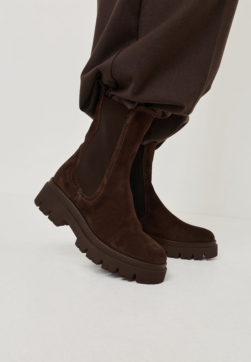 Bottines en daim marron avec semelles épaisses à crampons portées avec un pantalon marron assorti rentré à l'intérieur.