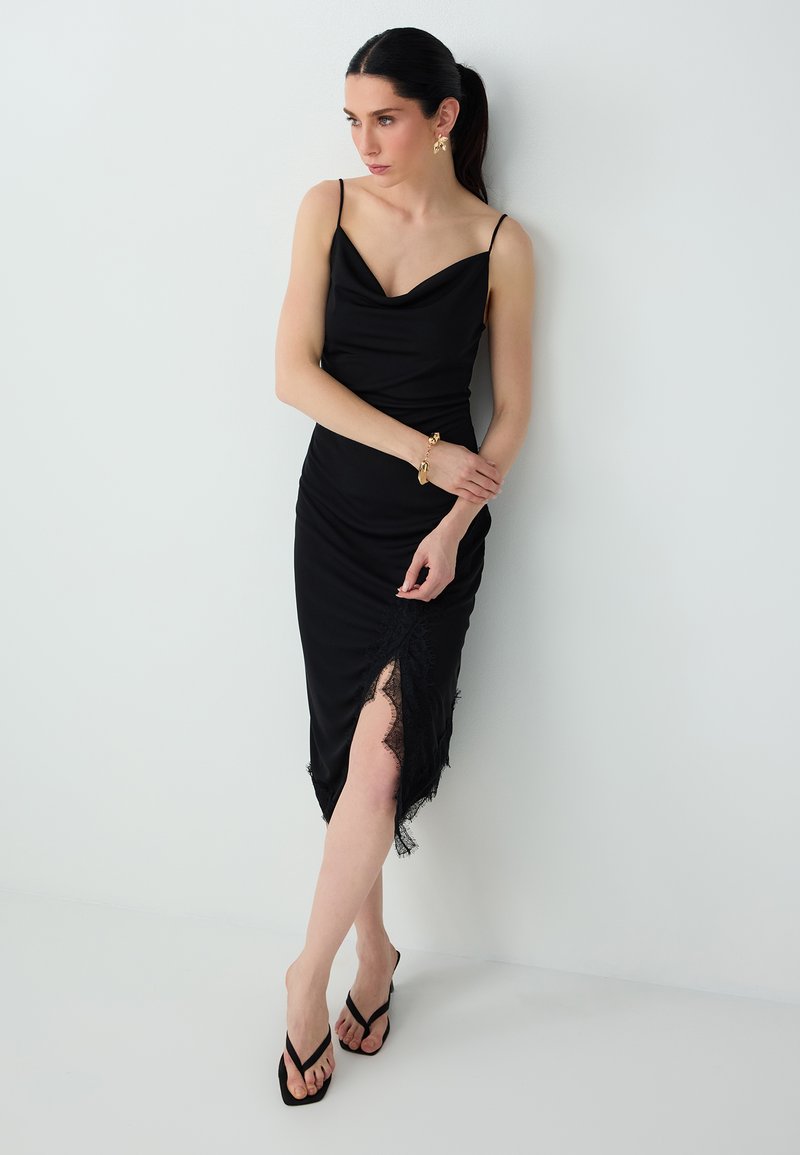 Femme en robe noire sans manches avec bordure en dentelle et fente, portant des bijoux dorés et des sandales noires, se tient devant un mur blanc uni.