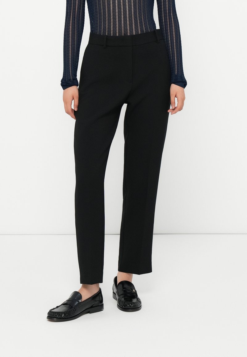 Femme portant un pantalon droit noir, des mocassins noirs brillants et un haut rayé transparent à manches longues bleu marine, se tenant devant un fond blanc.