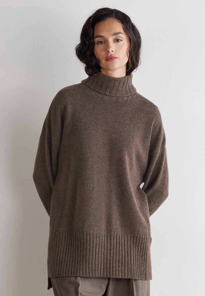 Femme aux cheveux bruns ondulés portant un pull à col roulé marron ample, sur un fond blanc uni.