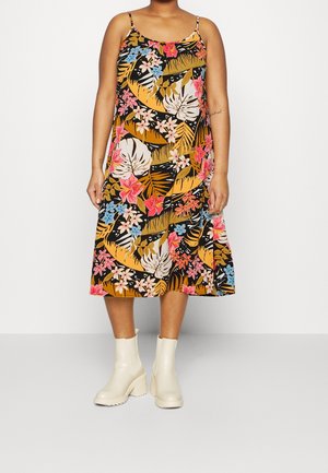 Vrouw draagt een mouwloze zwarte midi-jurk met een meerkleurige tropische bloemenprint en witte grove enkellaarzen, staand op een lichte vloer.