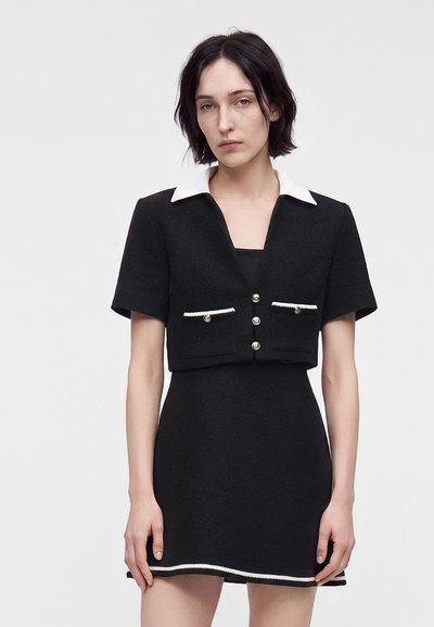 Femme aux cheveux courts et foncés portant une robe noire à manches courtes avec un col et des bordures blancs, debout devant un fond clair uni.