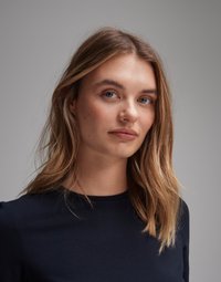 Portrait of a person with long, wavy, light brown hair, wearing a navy blue top with a round neckline, and neutral background.