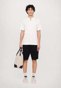 Polo shirt bej deschis cu guler cu fermoar, pantaloni scurți negri și adidași crem. Ținând o geantă bej cu accente negre. Fundal simplu.