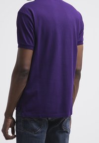 Lila bomullspolo, kortärmad, rund halsringning, visad bakifrån. Enkel design, inga synliga logotyper eller mönster, kombinerad med mörka jeans.