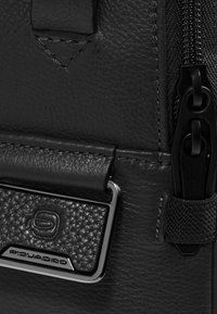 Detail einer schwarzen Ledertasche mit strukturierter Oberfläche, metallischen Beschlägen und einem Reißverschluss. Das Markenlogo ist auf einer glänzenden Metallplatte geprägt.