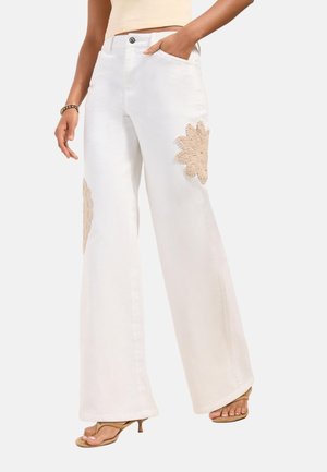 Persona che indossa pantaloni bianchi ampi con toppe floreali beige, sandali beige con tacco e top corto beige, mano in tasca.