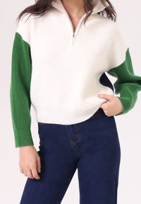 Suéter blanco acanalado con cremallera de cuarto de vuelta, que presenta mangas largas verdes y acentos laterales en azul marino, combinado con jeans azul oscuro.