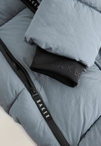 Doudoune grise avec texture matelassée, présentant un poignet élastique noir et un détail de fermeture éclair. L'étiquette indique "BAKER" en blanc.